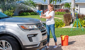 Woman using the Aqua Joe Fireman's Nozzle and hose to wash a car with an Aqua Joe sprinkler in the background watering the lawn