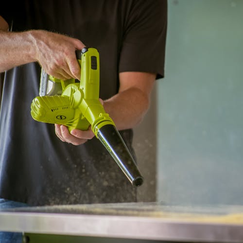 Person using the Sun Joe 24-volt cordless workshop blower with a 2.0-Ah lithium-ion battery attached to blow dust off a table.