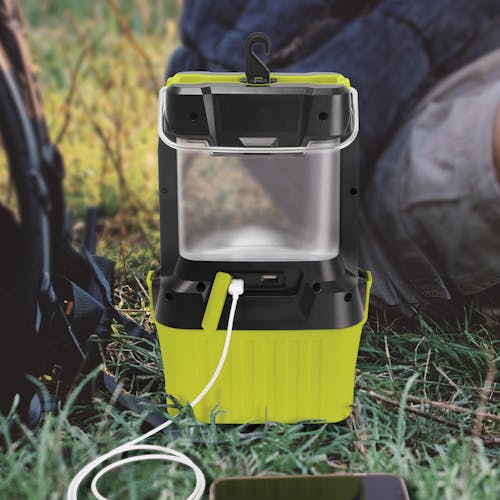 Sun Joe 24-Volt Cordless LED Lantern sitting in the grass and charging a cell phone.