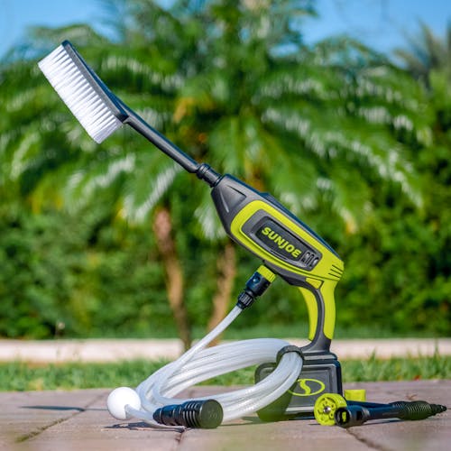 Sun Joe 24-Volt Cordless Power Cleaner outside on the ground with the brush attached and a palm tree in the background.