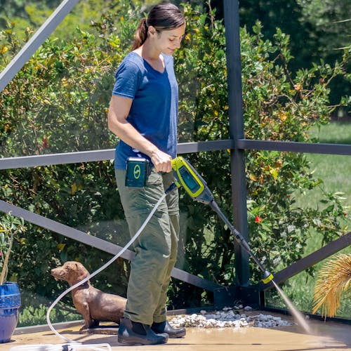 Woman using her Sun Joe 24-Volt Cordless Power Cleaner to clean her patio. Her small dog is behind her.