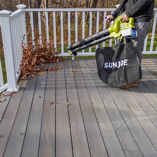 Person using the Sun Joe 48-volt cordless leaf blower, vacuum, mulcher with wheels with two 4.0-Ah lithium-ion batteries attached to blow leaves off a patio.