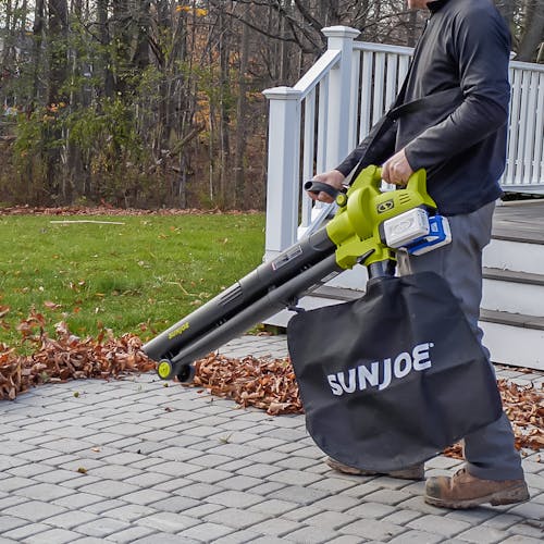 Person using the Sun Joe 48-volt cordless leaf blower, vacuum, mulcher with wheels with two 4.0-Ah lithium-ion batteries attached to blow leaves off paving stones in a backyard.