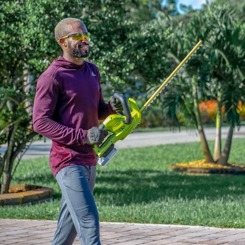 Man walking in his driveway holding the Side view of the Sun Joe 24-volt 22-inch cordless hedge trimmer with a 2.0-Ah lithium-ion battery attached.