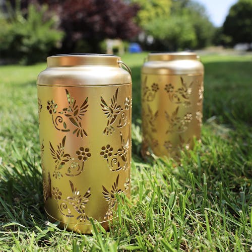 Set of 2 Gold Solar Lanterns with Humming Bird Design standing on grass.