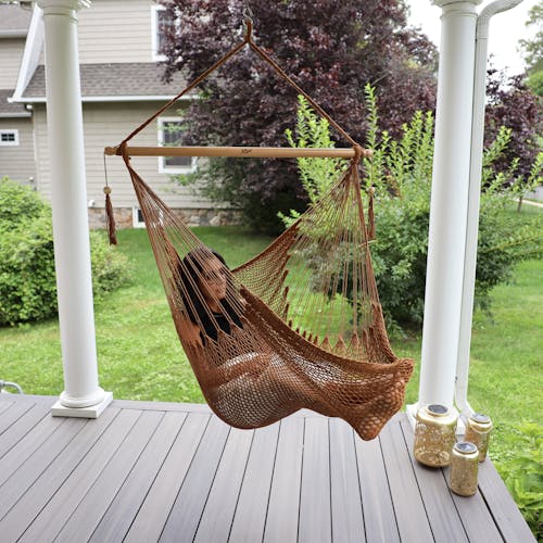 Woman sitting in the Bliss Hammocks 40-inch Brown Island Rope Hammock Chair hanging on a front porch.