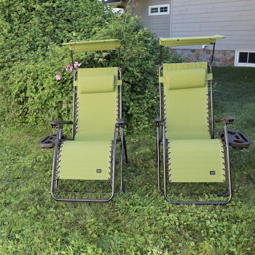 Set of 2 26-inch sage green gravity free chairs placed on a lawn in front of a lush green bush.