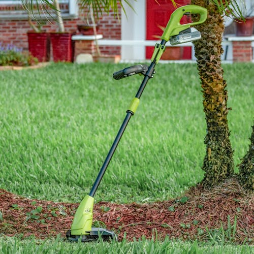 Sun Joe 24-volt cordless 10-inch stringless grass trimmer with a 2.0-Ah lithium-ion battery leaning against a tree.