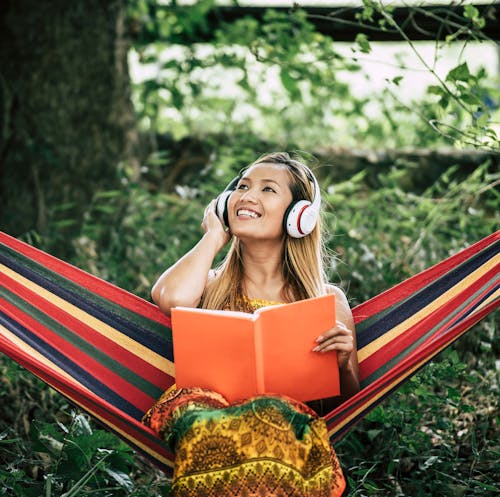 Woman with headphones on and a book in her hand sitting on a hammock outside.