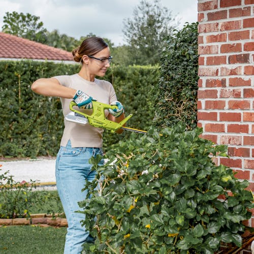 Woman using the Sun Joe 24-volt Cordless 18-inch hedge trimmer to trim a bush next to a house.