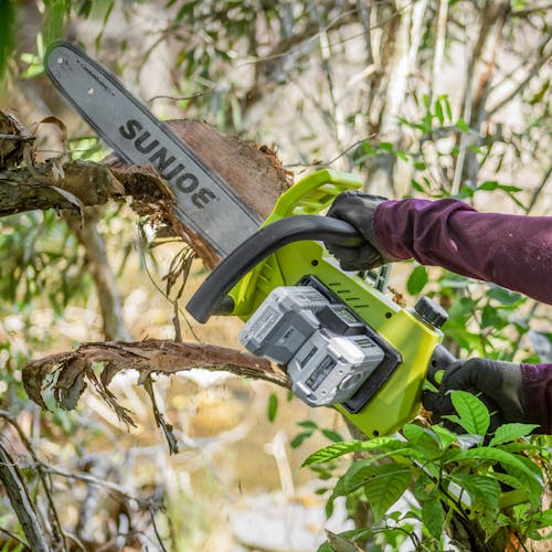 Person using the Sun Joe 48-volt cordless 16-inch chainsaw kit with two 2.0-Ah lithium-ion batteries attached to cut a branch off a tree.