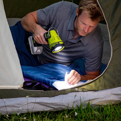 Man using the Sun Joe 24-Volt Cordless Flashlight/Flood Light/Spot Light to read a book inside a camping tent.