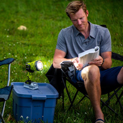 Man using the Sun Joe 24-Volt Cordless Flashlight/Flood Light/Spot Light to read a book outside while sitting in a camping chair.
