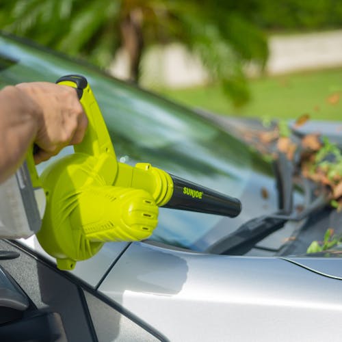 Person using the Sun Joe 24-volt cordless workshop blower with a 2.0-Ah lithium-ion battery attached to blow leaves off a car windshield.