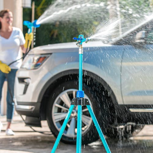 Aqua Joe 72-inch Indestructible Zinc Impulse 360-Degree Telescoping Tripod Sprinkler spraying water while a woman washes a car in the background.