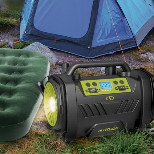 Sun Joe Hybrid 12-Volt/110-Volt AC High Volume Tire Inflator/Deflator on a camp with its headlight on next to an air mattress and camping tent.