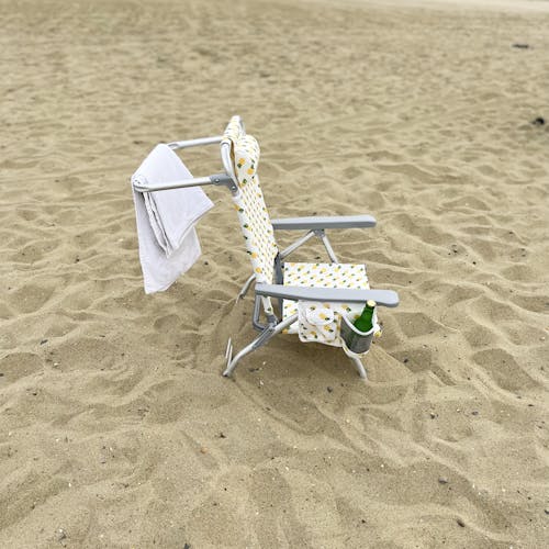 Side view of the folding pineapple beach chair on the sand with a towel hanging from the bar on the back and a drink in the side pocket.