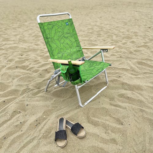 Angled view of the foldable green banana leaves beach chair on the sand with sandals next to it and a drink in the side pocket.