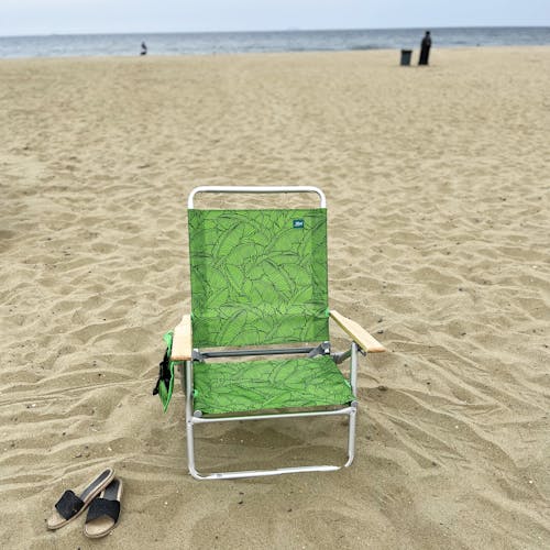 Front view of the foldable green banana leaves beach chair on the sand with sandals next to it.