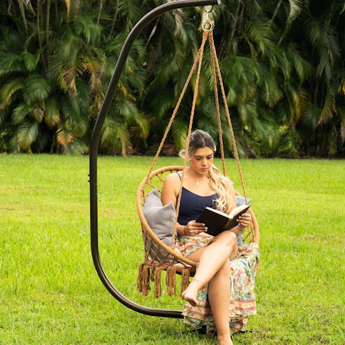 Woman reading a book while sitting in the Bliss Hammocks 31.5-inch Wide Brown Macramé Swing Chair outside in grass.