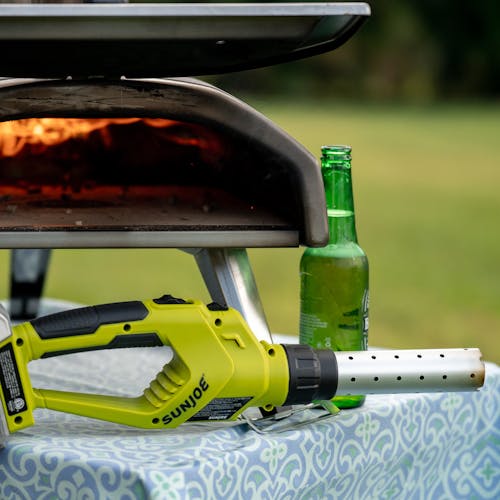 Sun Joe 24-Volt cordless electric fire starter and barbeque lighter laying on a table in front of a lit grill and a beer bottle.