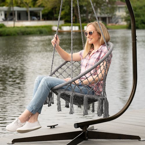 Woman sitting in the Bliss Hammocks 31.5-inch Wide Gray Macramé Swing Chair next to a river.