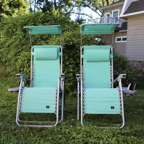 Set of 2 26-inch teal genome gravity free chairs on a lawn in front of a lush green bush.