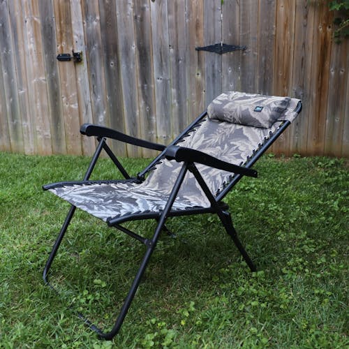 Angled view of a reclined 26-inch Platinum Fern Reclining Sling Chair on a lawn next to a fence.