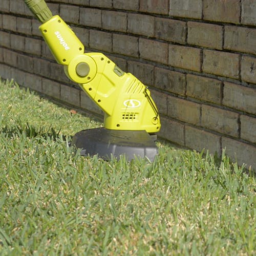 Sun Joe Electric Lawn Care System with the grass trimmer head being used to cut grass next to a brick wall.