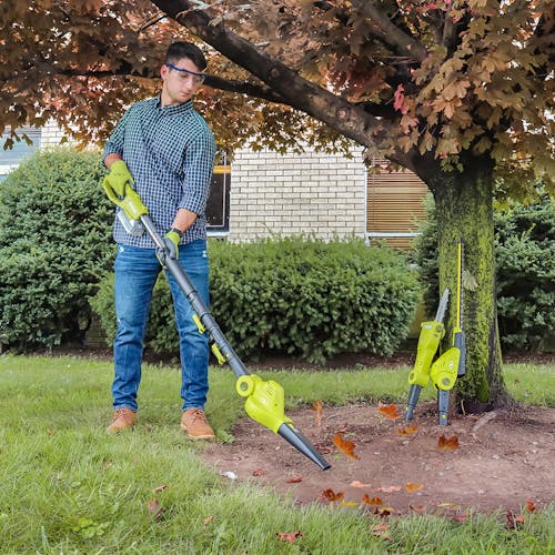 Man using the leaf blower attachment to blow leaves with the other two attachments leaning against a tree.