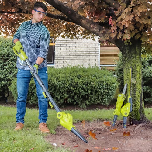 Man using the leaf blower attachment to blow leaves away from the ground around a tree. The pole saw and hedge trimmer attachments are leaning on the tree.