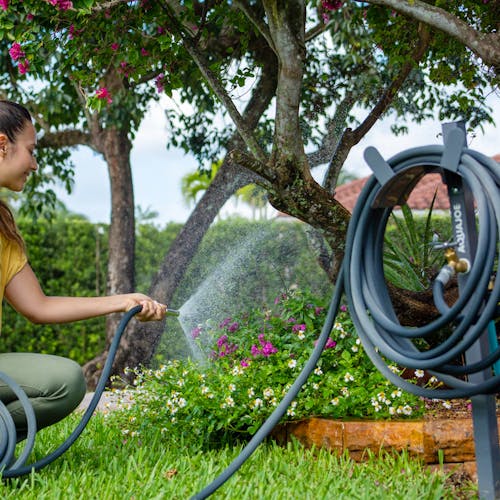 Aqua Joe Gray Garden Hose Stand with Brass Faucet holding a garden hose while a woman waters flowers.