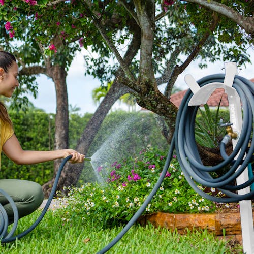 Aqua Joe White Garden Hose Stand with Brass Faucet holding a hose while a woman waters flowers.