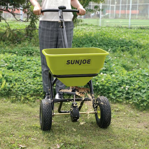 Sun Joe Multi-Purpose Walk-Behind Spreader being used to spread fertilizer on a lawn,