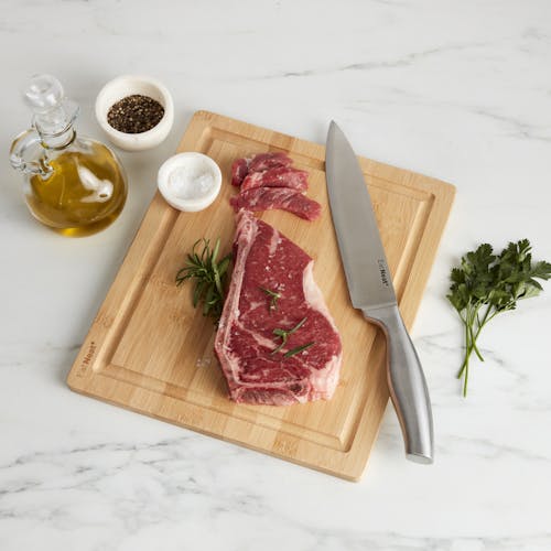 Cutting board for the EatNeat 8-piece knife set with a knife, steak, and spices on it with oil next to it on the kitchen counter.
