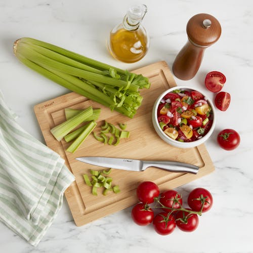 Cutting board for the EatNeat 8-piece knife set with a knife, tomatoes, and celery on it with a towel, oil and pepper next to it on the kitchen counter.
