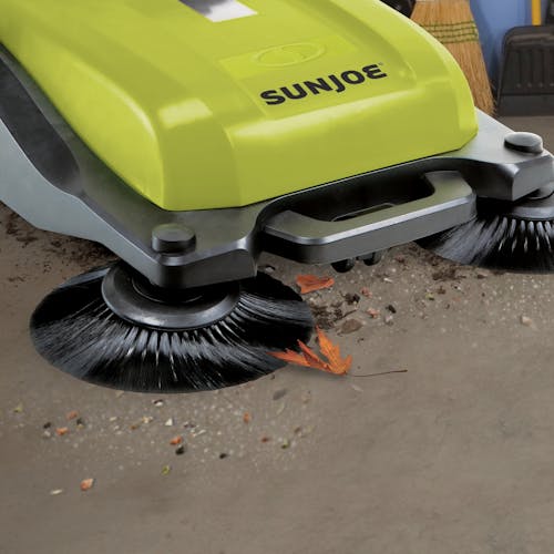 Close-up of the Sun Joe Push Driven Twin Brush Hard Surface Debris Sweeper sweeping leaves and debris off a floor.