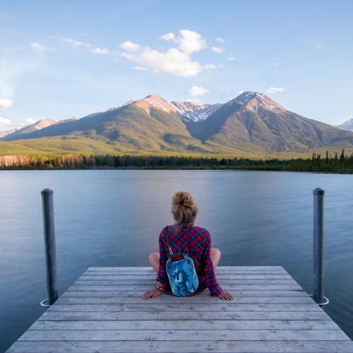 Woman sitting on a dock wearing a TrailGear blue camo dry bag with a lake and mountains in the background.