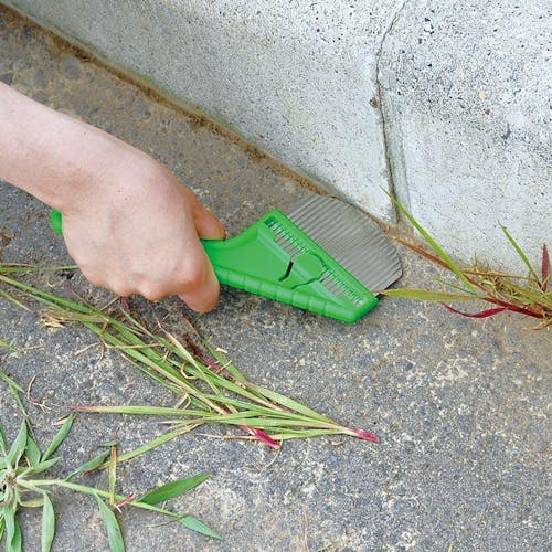 Nisaku Zassou Burush 3-inch Stainless Steel Rounded Weeder being used to get rid of weeds between the ground and a wall.